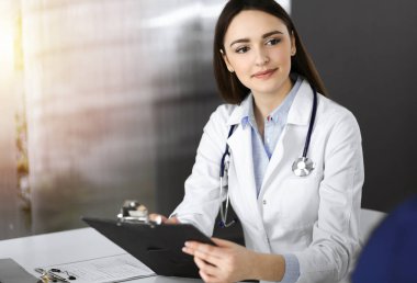 Dost canlısı genç bayan doktor, güneşli klinikte birlikte otururken hastasını dinliyor. Tıp ve sağlık hizmetleri kavramları