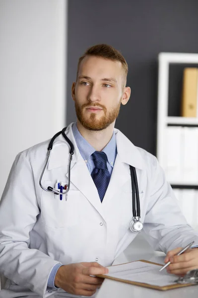 Vriendelijke rode baard dokter zittend en schrijvend aan klembord in kliniek. Perfecte medische dienst in het ziekenhuis. Geneesmiddelenconcept — Stockfoto