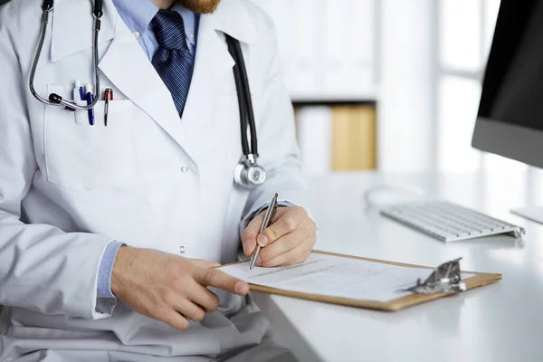 Amigável médico de barba vermelha sentado e escrevendo com área de transferência na clínica, close-up. Conceito de medicina — Fotografia de Stock