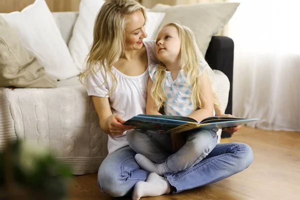 Glückliche Familie. Blonde junge Mutter liest ihrer süßen Tochter ein Buch vor, während sie am Holzboden sitzt. Mutterschaftskonzept — Stockfoto