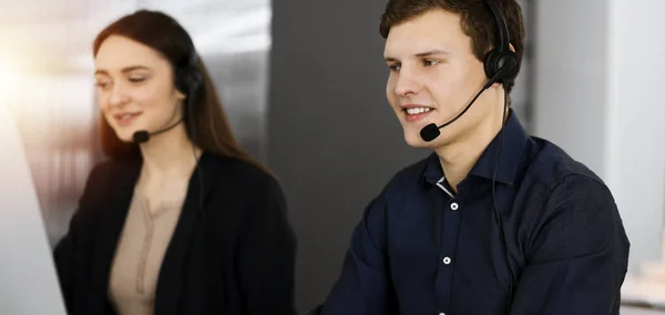Operadores de centro de llamadas en el trabajo. Dos jóvenes con auriculares están hablando con los clientes, mientras están sentados en una oficina soleada —  Fotos de Stock