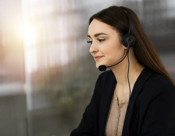 Jeune fille amicale dans les écouteurs parle à un client des entreprises, tout en étant assis au bureau ensoleillé. Opérateurs de centre d'appels au travail — Photo