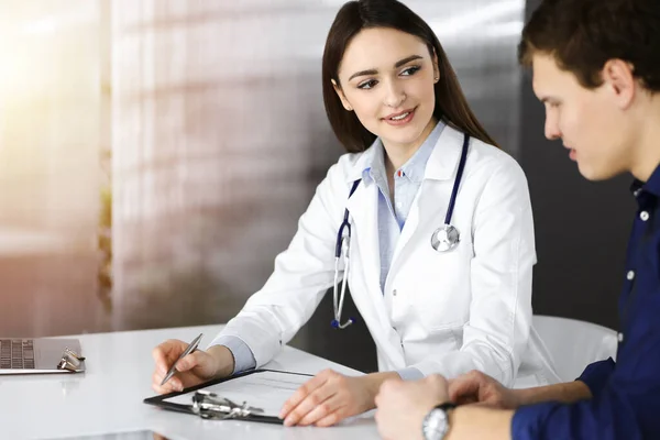 Freundliche junge Ärztin hört ihrer Patientin zu, während sie zusammen in der sonnigen Klinik sitzt. Medizin- und Gesundheitskonzepte — Stockfoto