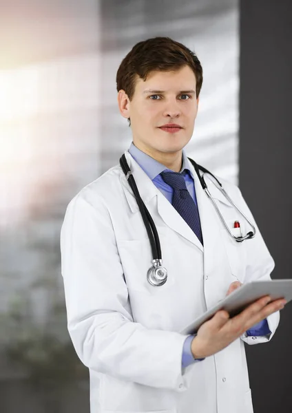 Ein junger freundlicher Arzt checkt ein paar Informationen auf seinem Computer-Tablet. Porträt eines Berufsarztes bei der Arbeit in einer sonnigen Klinik — Stockfoto
