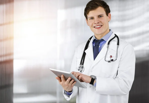 若いフレンドリーな医者は彼のコンピュータタブレットのいくつかの情報をチェックしている。日当たりの良いクリニックで働く専門医の肖像 — ストック写真