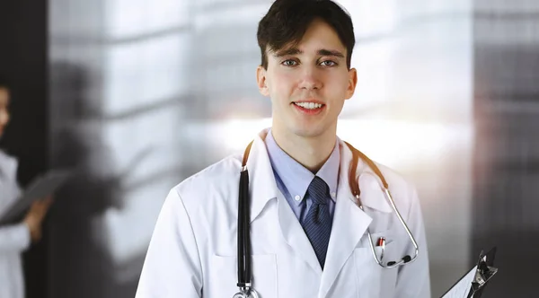 현대 의학에서 클립보드를 들고 서 있는 친절 한 젊은 남성 의사. 여성 동료는 의사의 배경으로 약학 기록을 작성하느라 바쁩니다. 의학적 개념 — 스톡 사진