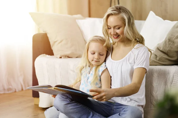 Bonne famille. Jeune mère blonde lisant un livre à sa jolie fille assise au sol en bois dans une pièce ensoleillée. Concept de maternité — Photo