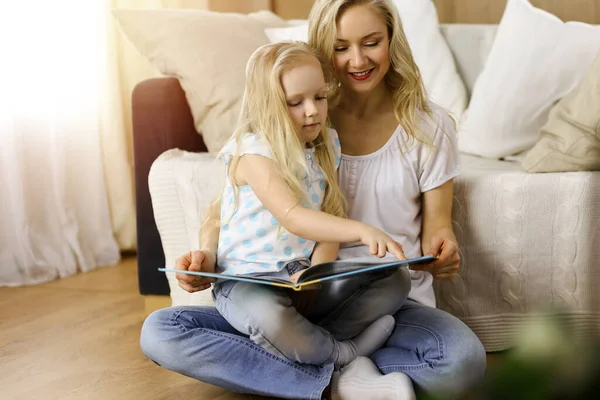 Glückliche Familie. Blonde junge Mutter liest ihrer süßen Tochter ein Buch vor, während sie am Holzboden in einem sonnigen Raum sitzt. Mutterschaftskonzept — Stockfoto