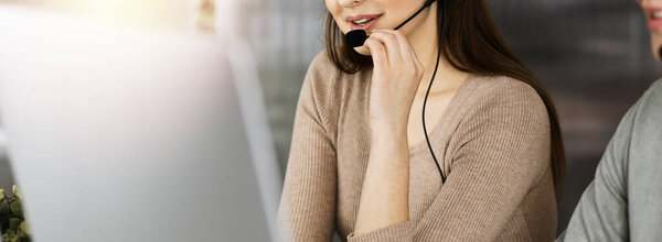 Two smiling people in headsets are talking to the clients, while sitting at the desk in sunny modern office