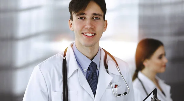 Vriendelijke jonge mannelijke arts staat met klembord in moderne kliniek. Vrouwelijke collega is bezig met het invullen van medicatiegegevens formulier op de achtergrond van de arts. Geneesmiddelenconcept — Stockfoto