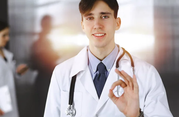 Freundlicher Mann-Arzt, der gerade steht und in der Klinik Ok-Zeichen zeigt. Die Kollegin hat es eilig im Hintergrund des Arztes. Medizinkonzept — Stockfoto