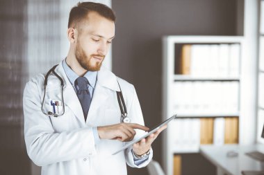 Dost canlısı kızıl sakallı doktor çalıştığı yerdeki klinikte tablet bilgisayar kullanıyor. Hastane ve tıp konseptinde mükemmel bir tıbbi hizmet.