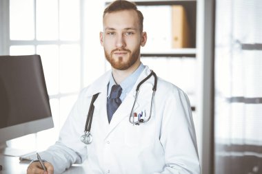 Dost canlısı kızıl sakallı doktor klinikteki panoda oturuyor ve yazıyor. Hastanede mükemmel bir tıbbi hizmet. Tıp konsepti