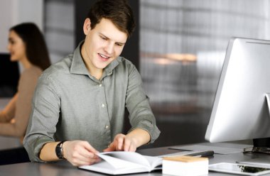 Yeşil gömlekli genç işadamı ve programcı arka planda bir bayan meslektaşıyla birlikte modern bir dolapta otururken defterini çeviriyor ve kameraya bakıyor.