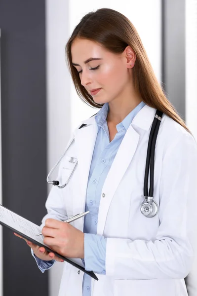 Medico donna riempiendo modulo medico mentre in piedi vicino alla finestra in ufficio ospedaliero. Medico felice al lavoro. Medicina e concetto di assistenza sanitaria — Foto Stock