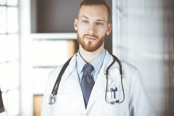 Κοκκινογένης γιατρός στέκεται ακριβώς στην κλινική κοντά στο χώρο εργασίας του. Πορτρέτο γιατρού. Ιατρική και υγειονομική περίθαλψη έννοια — Φωτογραφία Αρχείου