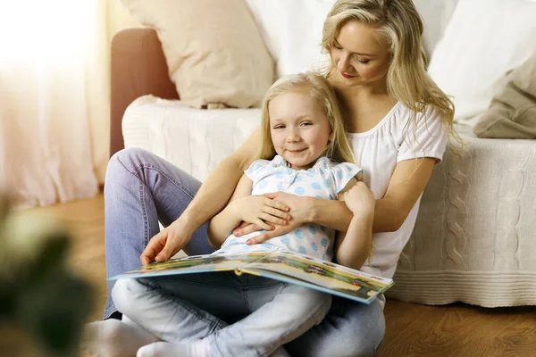 Bonne famille. Jeune mère blonde lisant un livre à sa jolie fille assise au sol en bois dans une pièce ensoleillée. Concept de maternité — Photo