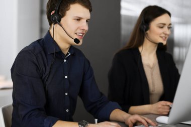 Modern bir ofiste masada otururken, kulaklıklı iki genç müşterilerle konuşuyor. Mavi gömlekli adama odaklan. İş yerindeki çağrı merkezi operatörleri