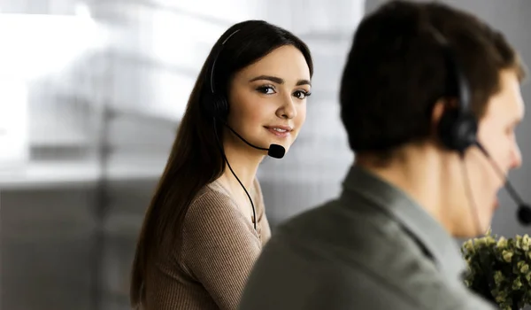 Joven chica alegre en auriculares está mirando a la cámara, mientras está sentado en el escritorio con un colega masculino. Retrato del operador del centro de llamadas en el trabajo —  Fotos de Stock