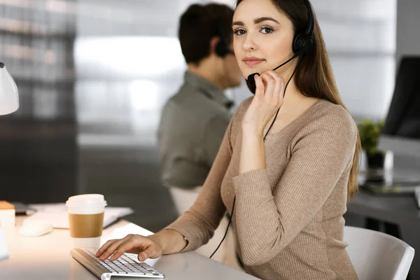 Jeune fille amicale dans les écouteurs parle à un client des entreprises, tout en étant assis sur le bureau dans un bureau moderne avec son collègue. Opérateurs de centre d'appels au travail — Photo