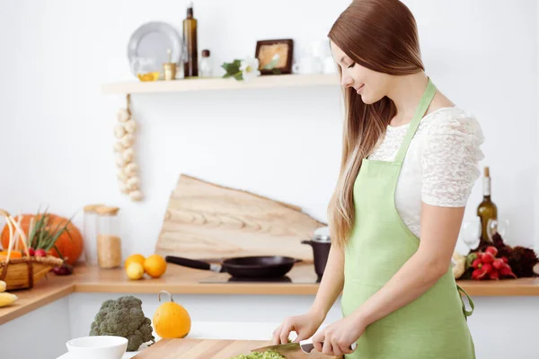 Όμορφη γυναίκα ντυμένη με ποδιά μαγειρεύει στην κουζίνα. Νοικοκυρά τεμαχίζει φρέσκια χορτοφαγική σαλάτα — Φωτογραφία Αρχείου