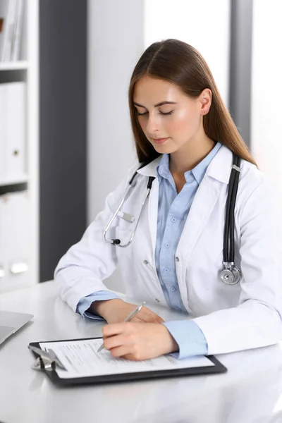 Orvos nő kitölti az orvosi papírokat, miközben az asztalnál ül a kórházi irodában. Orvos a munkahelyemen. Orvosi és egészségügyi ellátás fogalma — Stock Fotó