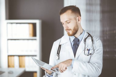 Dost canlısı kızıl sakallı doktor çalıştığı yerdeki klinikte tablet bilgisayar kullanıyor. Hastane ve tıp konseptinde mükemmel bir tıbbi hizmet.