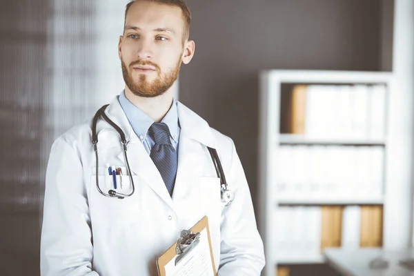 Red-bearded doctor standing straight in clinic near his working place. Portrait of physician. Medicine and healthcare concept