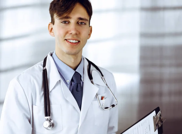 Barátságos fiatal férfi orvos áll írótáblával a modern klinikán. Orvosi fogalom — Stock Fotó