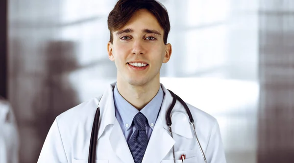 Amical jeune médecin masculin debout avec presse-papiers dans la clinique moderne. Une collègue féminine est occupée à remplir le formulaire de dossier médical à l'arrière-plan du médecin. Concept de médecine — Photo