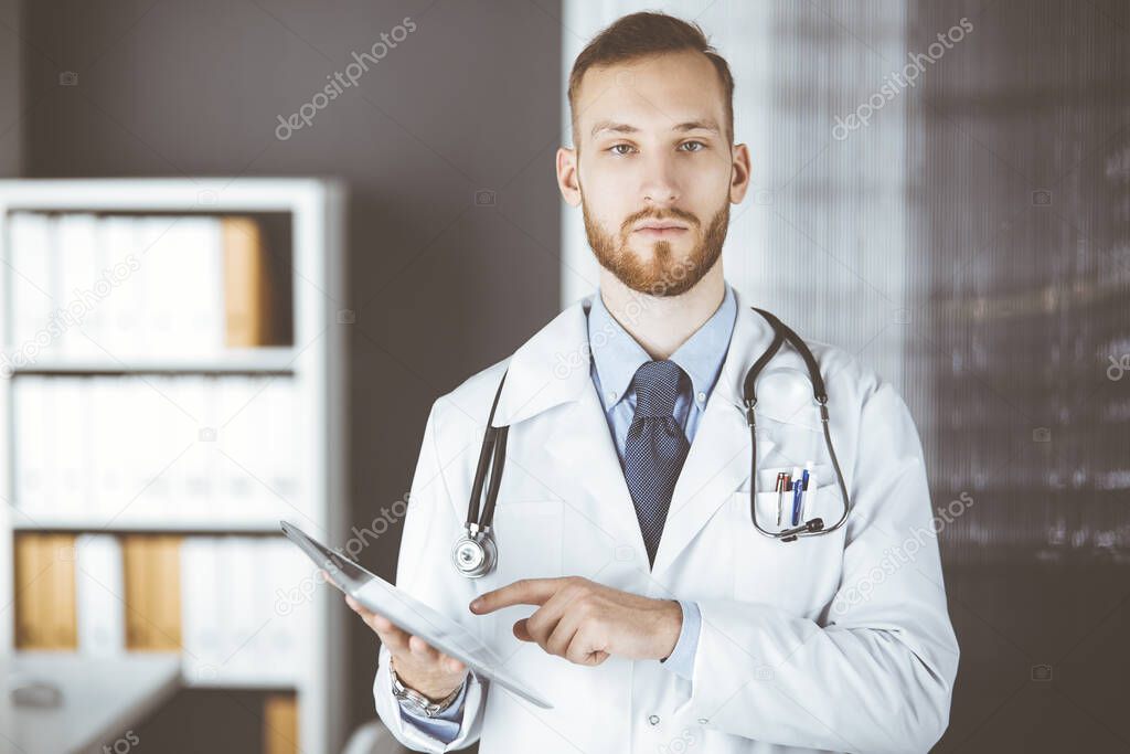 Friendly red-bearded doctor using tablet computer in clinic at his working place. Perfect medical service in hospital and medicine concept