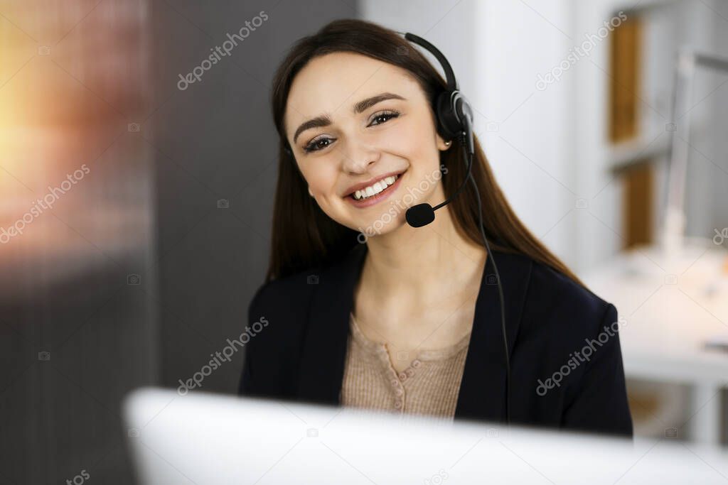 Young friendly girl in headsets is talking to a firms client, while sitting at the desk in sunny office. Call center operators at work