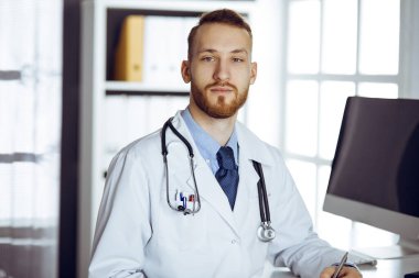 Dost canlısı kızıl sakallı doktor klinikteki panoda oturuyor ve yazıyor. Hastanede mükemmel bir tıbbi hizmet. Tıp konsepti