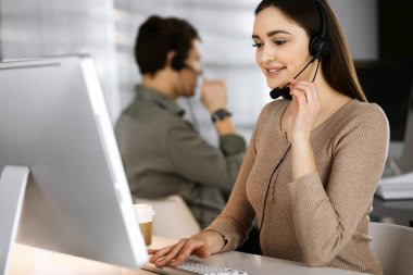 Modern bir ofiste meslektaşıyla birlikte masa başında otururken, kulaklıklı genç bir kız bir firma müşterisiyle konuşuyor. İş yerindeki çağrı merkezi operatörleri