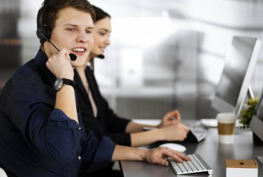 Modern bir ofiste masada otururken, kulaklıklı iki genç müşterilerle konuşuyor. Mavi gömlekli adama odaklan. İş yerindeki çağrı merkezi operatörleri