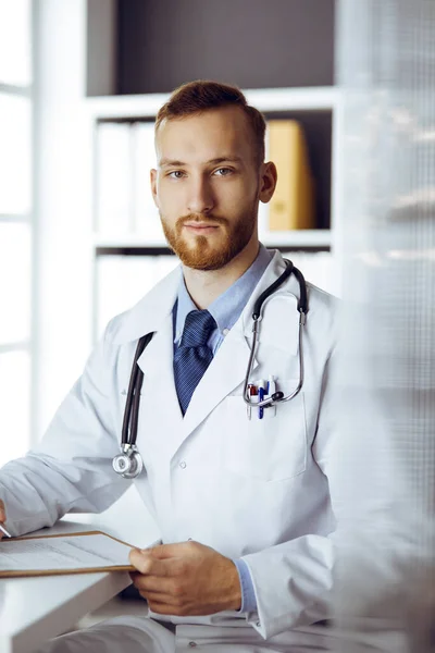 Amichevole medico barbuto seduto e scrivere negli appunti in clinica. Servizio medico perfetto in ospedale. Concetto di medicina — Foto Stock