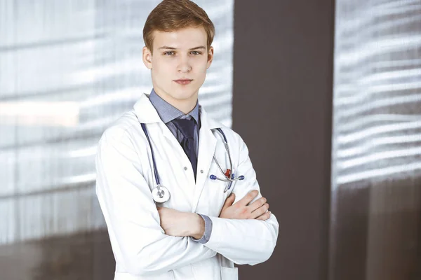 Jovem médico de pé com os braços cruzados na clínica perto de seu local de trabalho. Médico loiro no trabalho. Serviço médico perfeito no hospital. Conceito de medicina — Fotografia de Stock