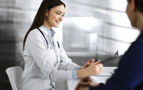 Une jeune femme-médecin amicale écoute sa patiente, alors qu'ils sont assis ensemble au bureau dans un cabinet. Médecin tient un presse-papiers dans ses mains pour remplir les médicaments — Photo