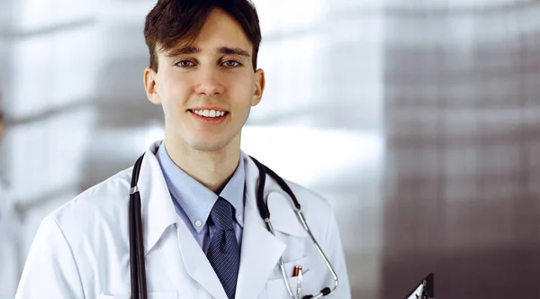 Φιλικός νεαρός άνδρας γιατρός στέκεται με πρόχειρο στη σύγχρονη κλινική. Γυναίκα συνάδελφος είναι απασχολημένος με τη συμπλήρωση της φόρμας αρχείων φαρμακευτική αγωγή στο παρασκήνιο του γιατρού. Έννοια ιατρικής — Φωτογραφία Αρχείου