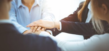 İş dünyası ekibi güneşli ofiste ellerini birleştiriyor. El ele tutuşan ve arkadaşlık, takım çalışması ve ortaklık kavramını temsil eden bir grup insan