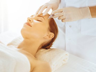 Güzel bir kadın güneşli tıp merkezinde kapalı gözlerle güzellik iğneleri yaptırıyor. Güzellik uzmanı elleri şırıngayla kadın yüzüne güzellik prosedürü uyguluyor.