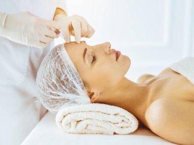 Güzel bir kadın güneşli tıp merkezinde kapalı gözlerle güzellik iğneleri yaptırıyor. Güzellik uzmanı elleri şırıngayla kadın yüzüne güzellik prosedürü uyguluyor.