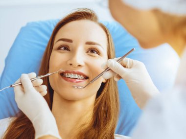 Güneşli diş kliniğinde dişçi tarafından incelenen diş telleriyle gülümseyen genç bir kadın. Sağlıklı diş ve ilaç konsepti