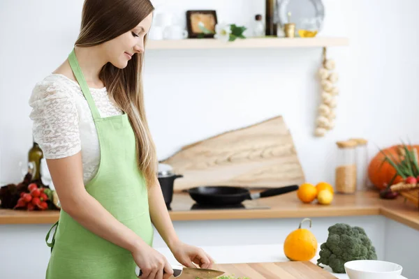 Hermosa mujer vestida de delantal está cocinando comida en la cocina. Ama de casa rebanando ensalada vegana fresca —  Fotos de Stock