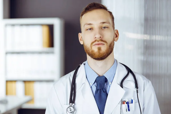 Red-bearded doctor standing straight in clinic near his working place. Portrait of physician. Medicine and healthcare concept