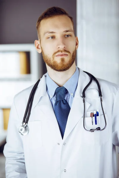 Kızıl sakallı doktor çalışma yerinin yanındaki klinikte dikiliyor. Hekim portresi. Tıp ve sağlık hizmetleri kavramı — Stok fotoğraf