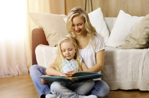 Bonne famille. Jeune mère blonde lisant un livre à sa jolie fille assise au sol en bois dans une pièce ensoleillée. Concept de maternité — Photo