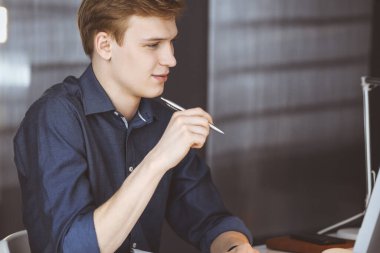 Genç sarışın işadamı iş yerinde bilgisayarla strateji düşünüyor. Girişim işi, başarı için çok çalışmak ve zamanın dışında çalışmak demektir.