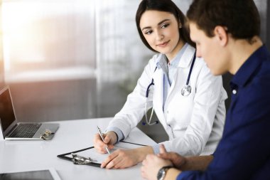 Dost canlısı genç bayan doktor, güneşli klinikte birlikte otururken hastasını dinliyor. Tıp ve sağlık hizmetleri kavramları