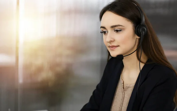 Jeune fille amicale dans les écouteurs parle à un client des entreprises, tout en étant assis au bureau ensoleillé. Opérateurs de centre d'appels au travail — Photo